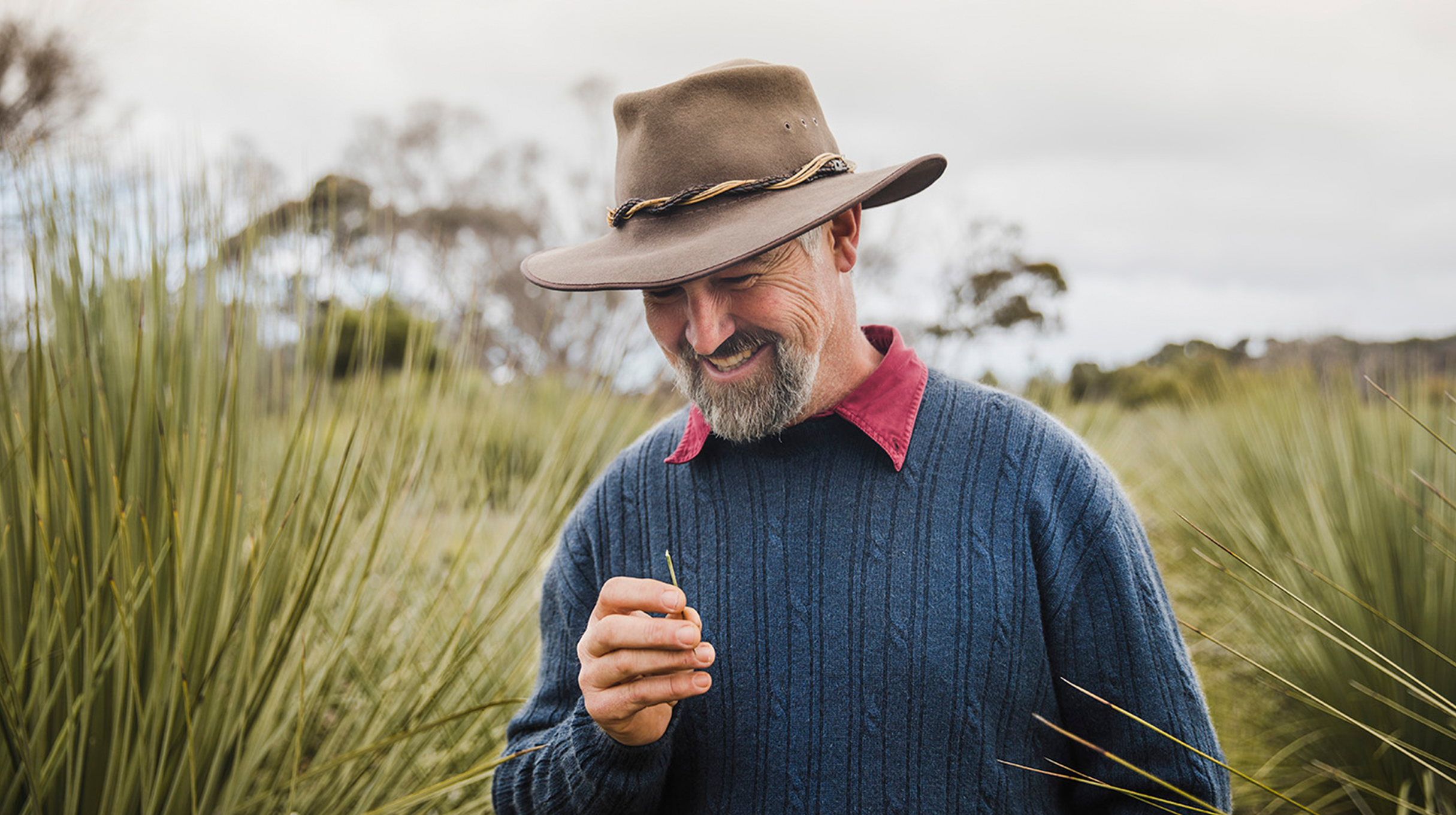 Middle-aged man looking down, holding a blade of grass, wearing an akubra-style hat, and a blue cable knitted woollen jumper.