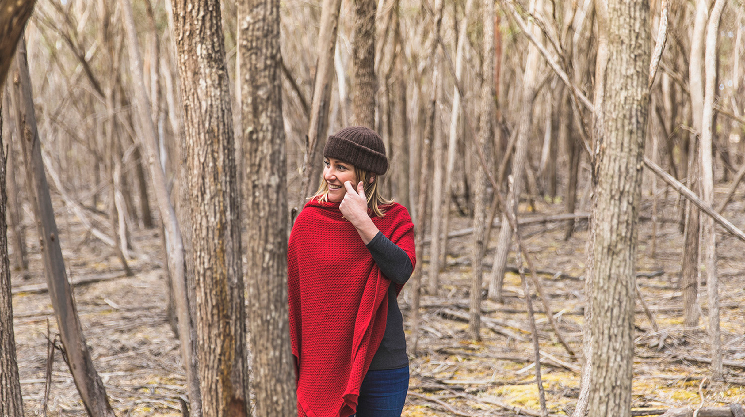Woman surrounded by tree trunks, wearing a bright red woollen poncho and brown woollen beanie.