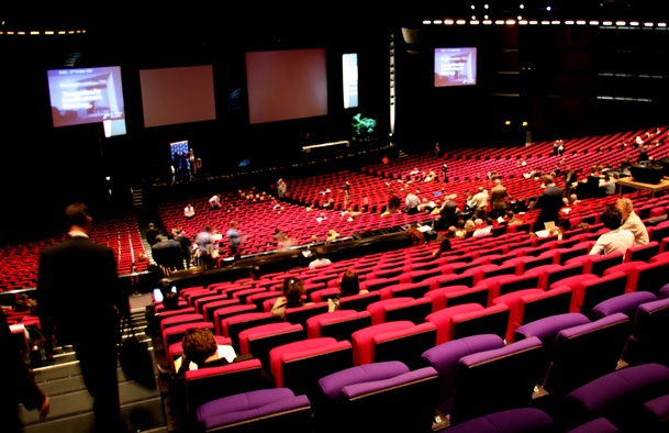 A theatre with red seats with people filing in to sit down