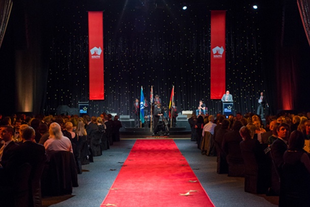 A red carpet leading to a stage in a theatre, people are sitting on either side. The South Australia logo is displayed on large banners on stage.