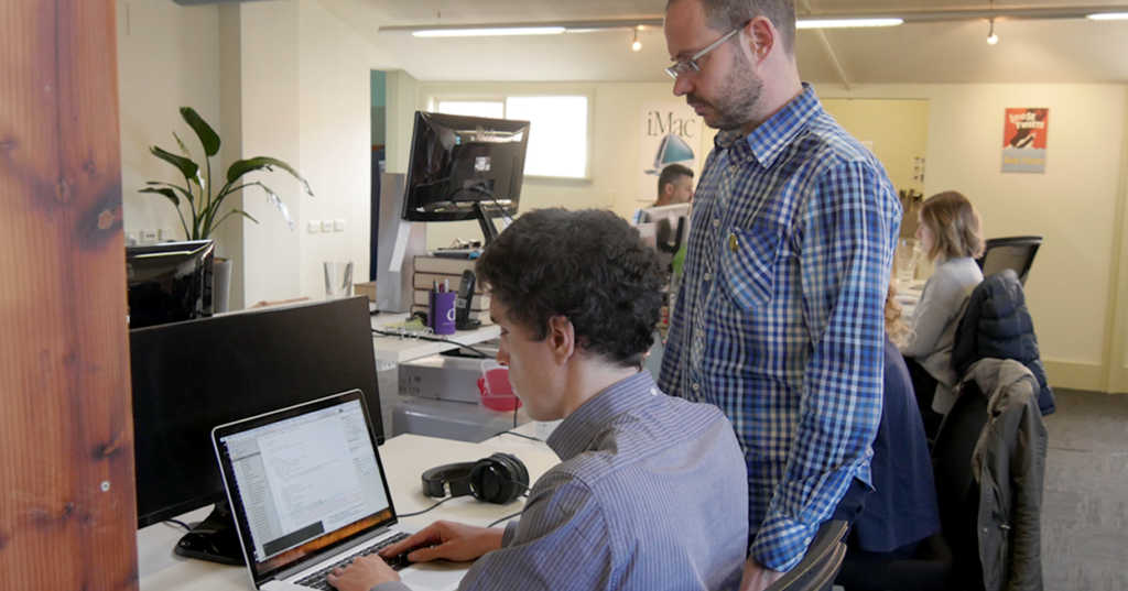 Tim is typing into a laptop. Nick is standing next to him and looking at the brilliant work that Tim is producing. 